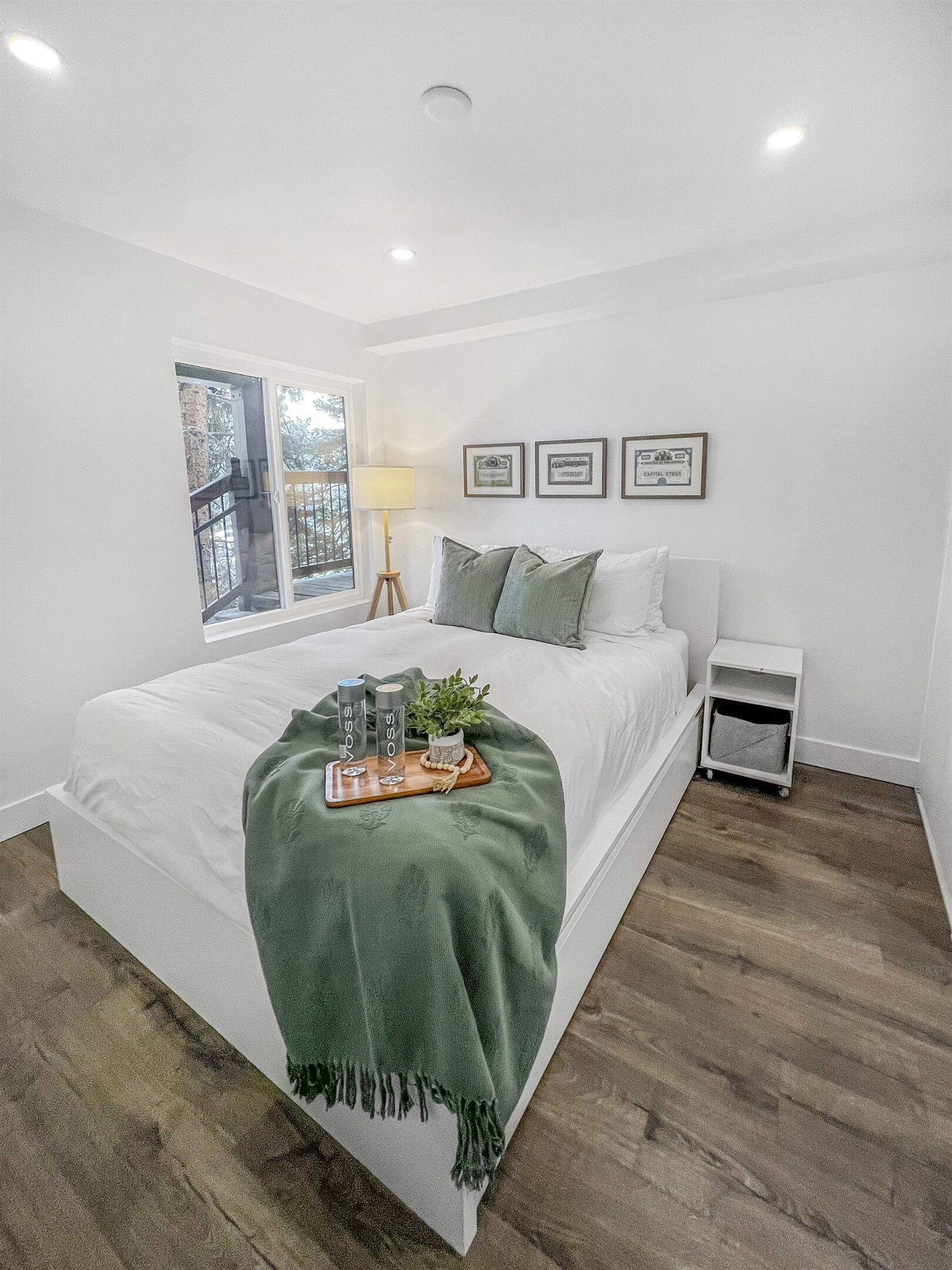 A bedroom with a bed, a bedside table, a lamp, a window, and framed pictures on the wall.