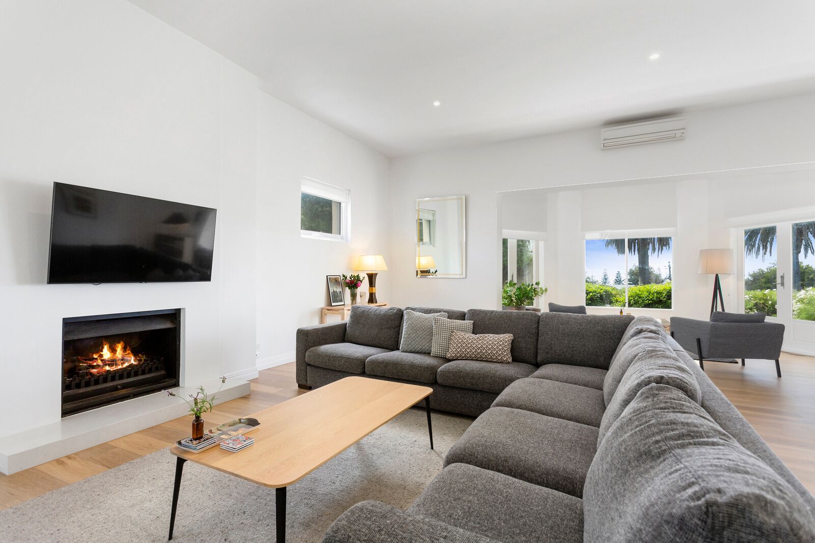 Una sala de estar moderna con chimenea, sofás grises, televisor y vistas a zonas verdes a través de grandes ventanales.