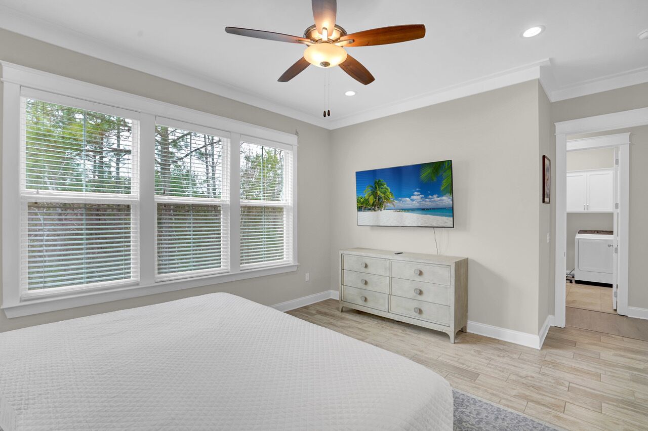 A bedroom with a bed, a ceiling fan, a painting, a dresser, and a laundry area.