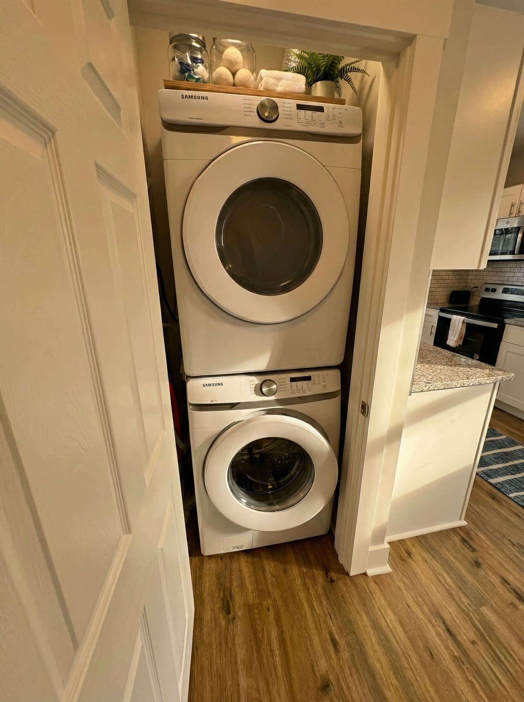 Une petite buanderie avec des lave-linge et des sèche-linge superposés, une petite étagère avec des articles de toilette et un coin cuisine visible à travers une porte ouverte.