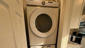 A compact laundry room with stacked washers and dryers, a small shelf with toiletries, and a kitchen area visible through an open door. - Newly Renovated Apt with King Bed (Wilmington)