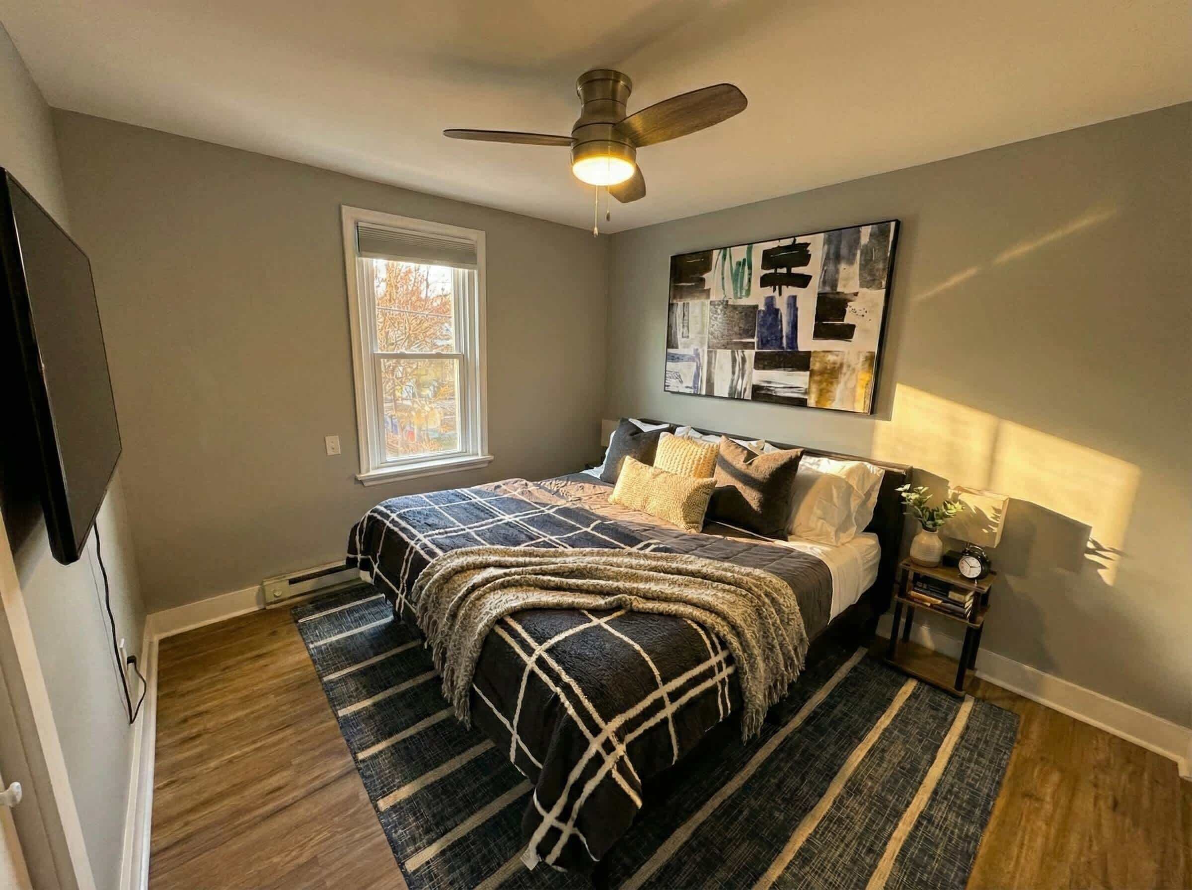 Une chambre à coucher comprenant un lit, un ventilateur de plafond, une fenêtre et une œuvre d’art accrochée au mur.