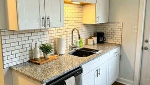 A compact kitchen with white cabinets, a granite countertop, a dishwasher, and a tiled backsplash. - Newly Renovated Apt with King Bed (Wilmington)