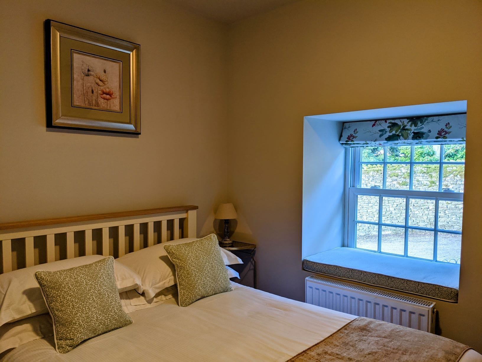 A bedroom with a bed, pillows, a nightstand, a lamp, a framed picture, and a window with a view of a stone wall and greenery.