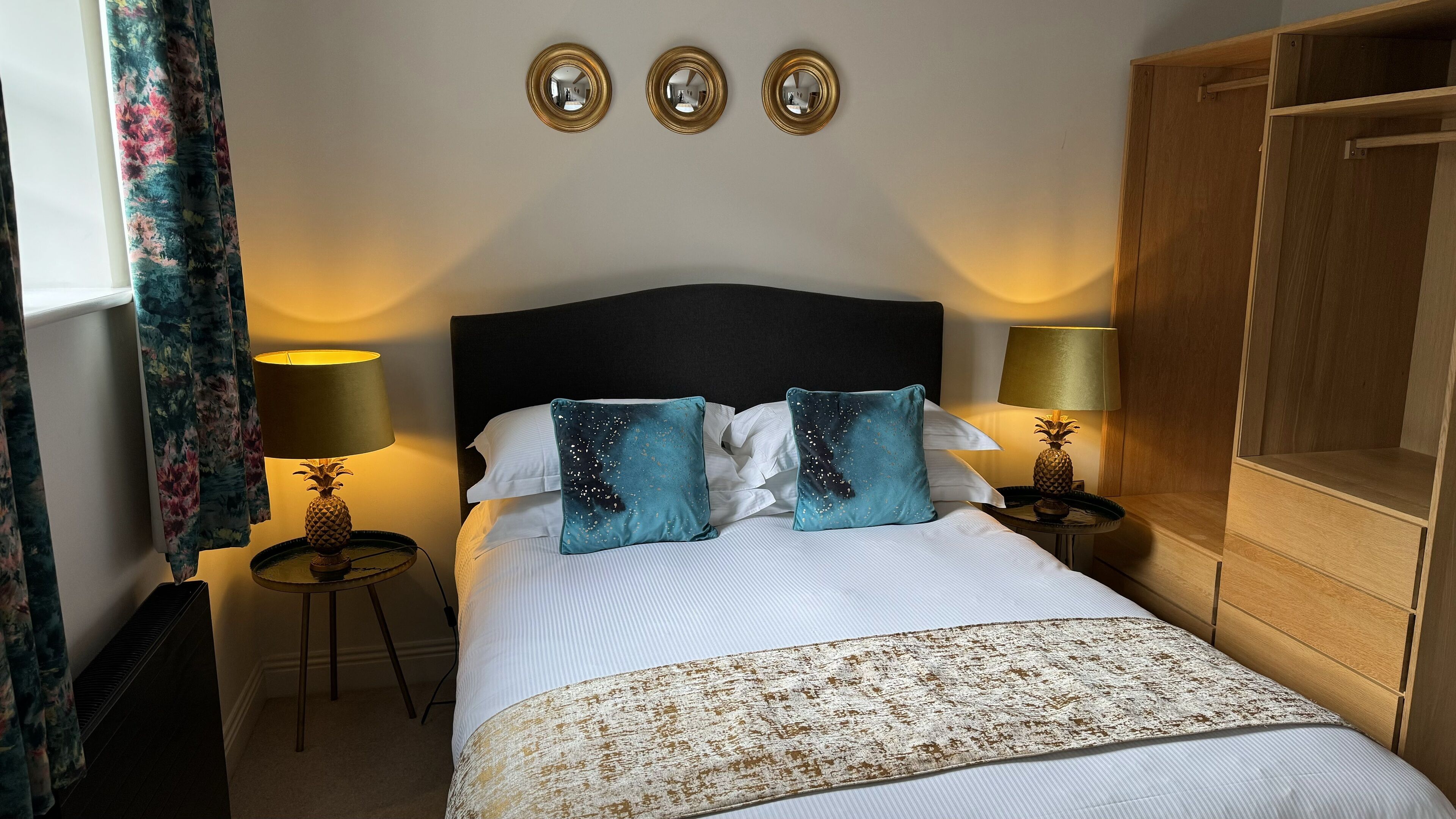 A neatly made bed with white linens, two teal pillows, and a patterned throw. There are two bedside tables with lamps, one of which has a pineapple decoration. The room has a dark headboard, a wooden wardrobe, and three wall-mounted mirrors.