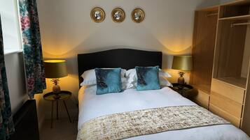 A neatly made bed with white linens, two teal pillows, and a patterned throw. There are two bedside tables with lamps, one of which has a pineapple decoration. The room has a dark headboard, a wooden wardrobe, and three wall-mounted mirrors.
