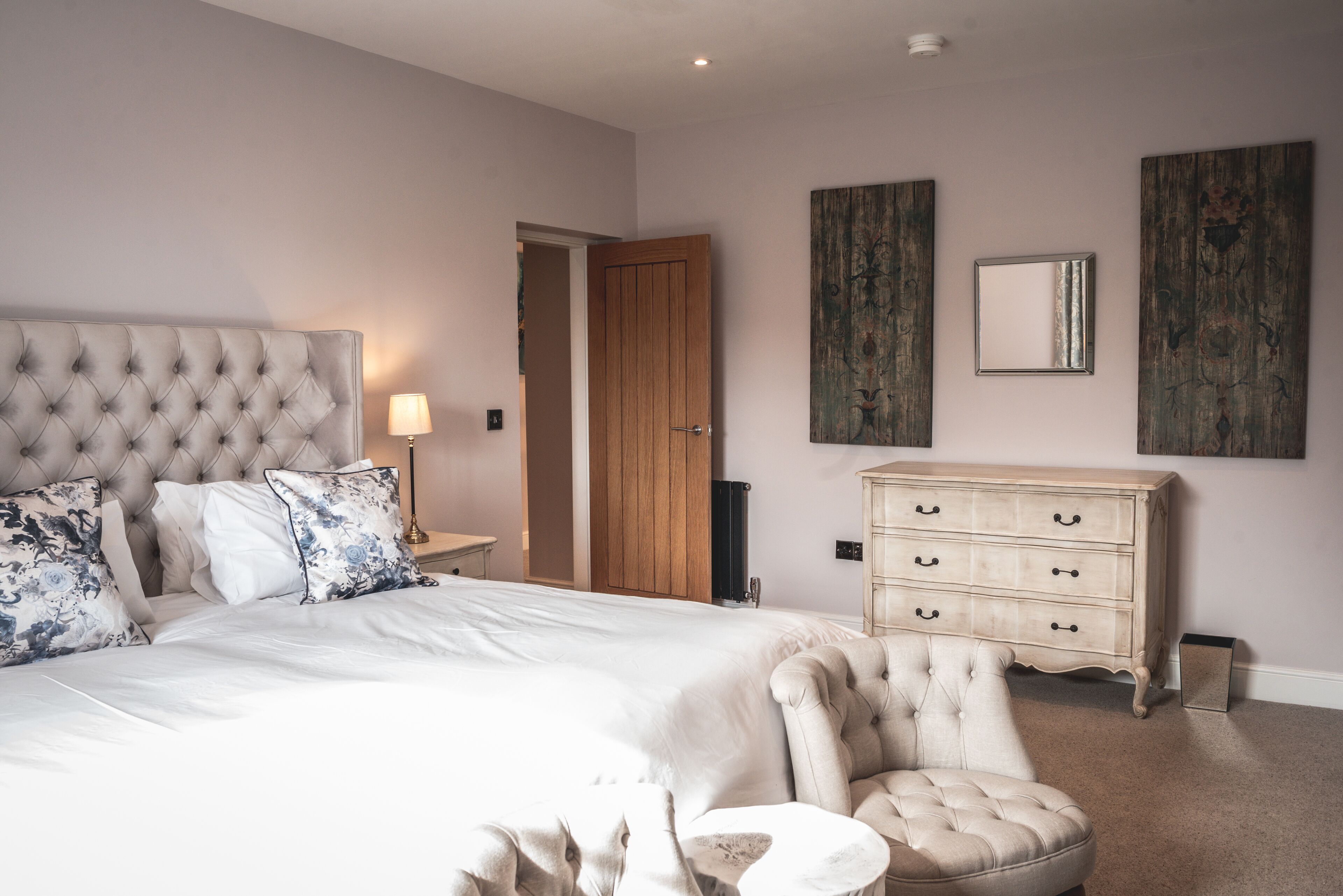 A bedroom with a tufted headboard, a large bed with white linens, a cream-colored armchair, a wooden dresser, and two wall-mounted mirrors.