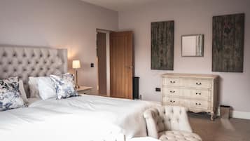 Ein Schlafzimmer mit einem gepolsterten Kopfteil, einem großen Bett mit weißen Leinen, einem cremefarbenen Sessel, einer Holzkommode und zwei Wandspiegeln.