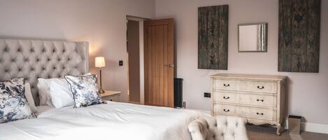 A bedroom with a tufted headboard, a large bed with white linens, a cream-colored armchair, a wooden dresser, and two wall-mounted mirrors.