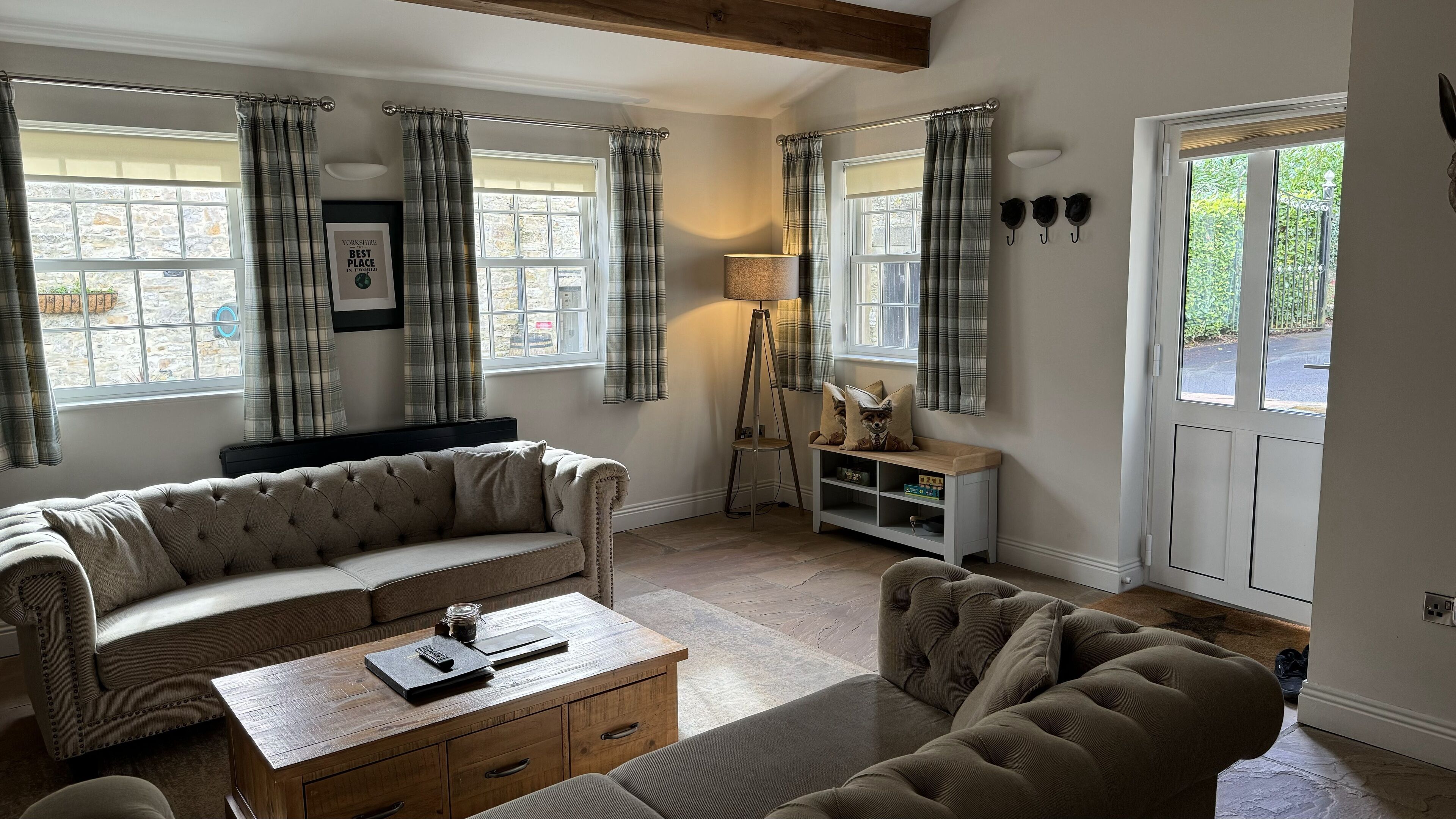 A cozy living room with a sofa, a coffee table, a lamp, and a side table. There is a window with curtains, a door, and a small shelf with decorative items.