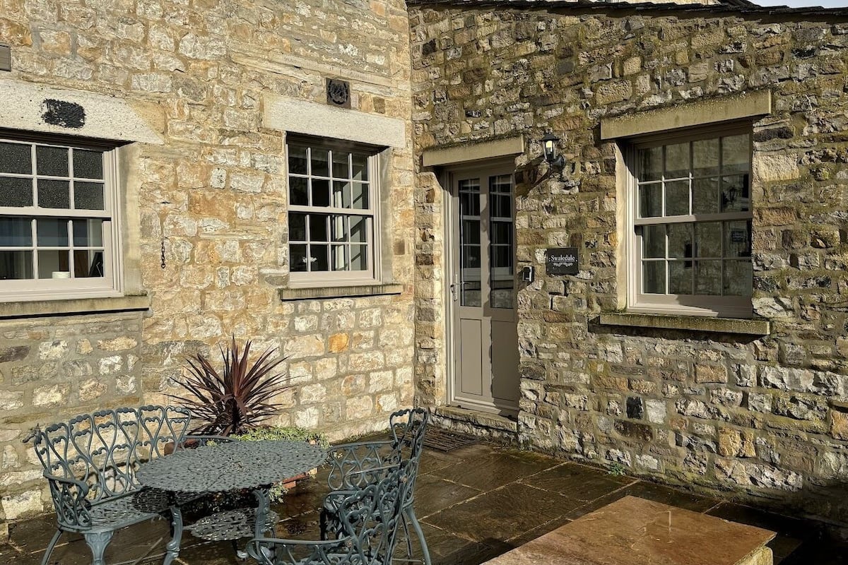 Edificio de piedra con un pequeño patio amueblado con mesa y sillas de metal, una puerta y una ventana con un cartel.