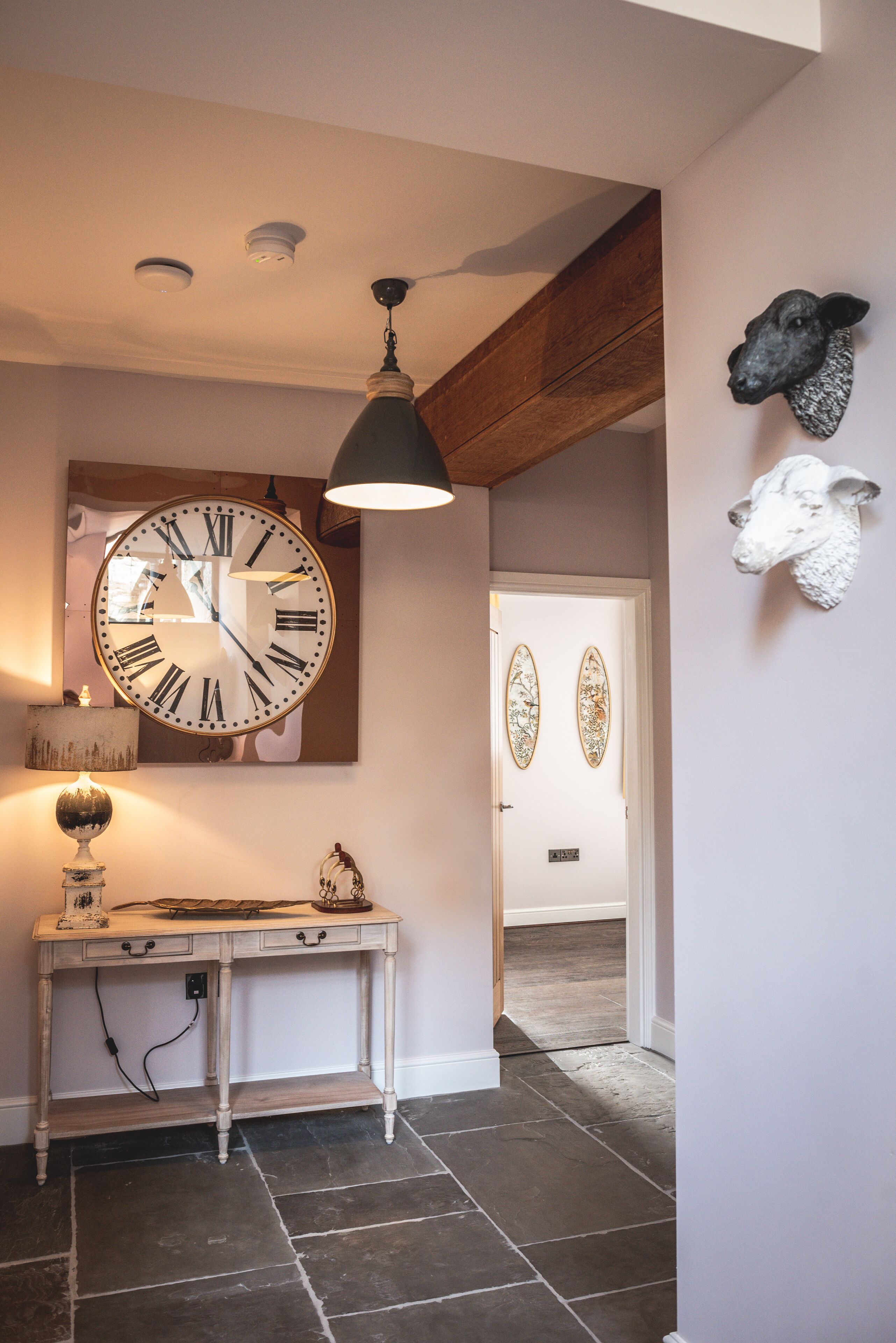 A hallway with a large wall clock, a wooden ceiling beam, and decorative sheep heads on the wall.