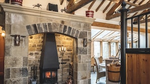 A stone fireplace with a black mantle, a wooden ceiling with exposed beams, and a wicker chair near the fireplace. - The Saddle Room Restaurant (Leyburn)