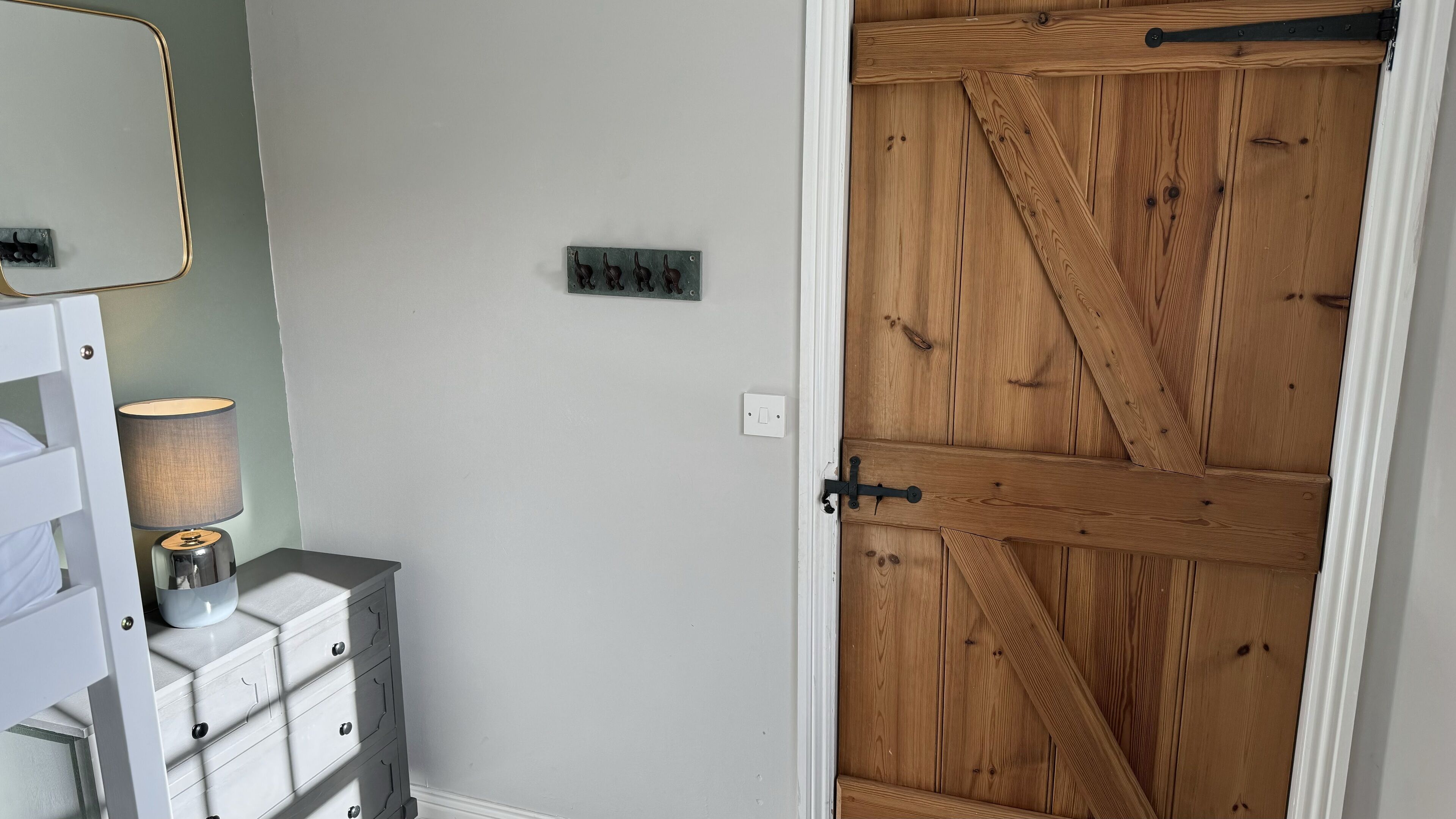 A room with a white dresser, a lamp, a wall-mounted light switch, and a wooden door with a metal latch.