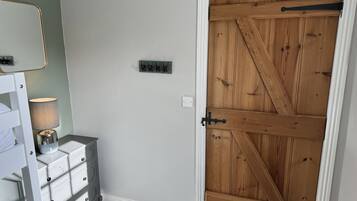 A room with a white dresser, a lamp, a wall-mounted light switch, and a wooden door with a metal latch.
