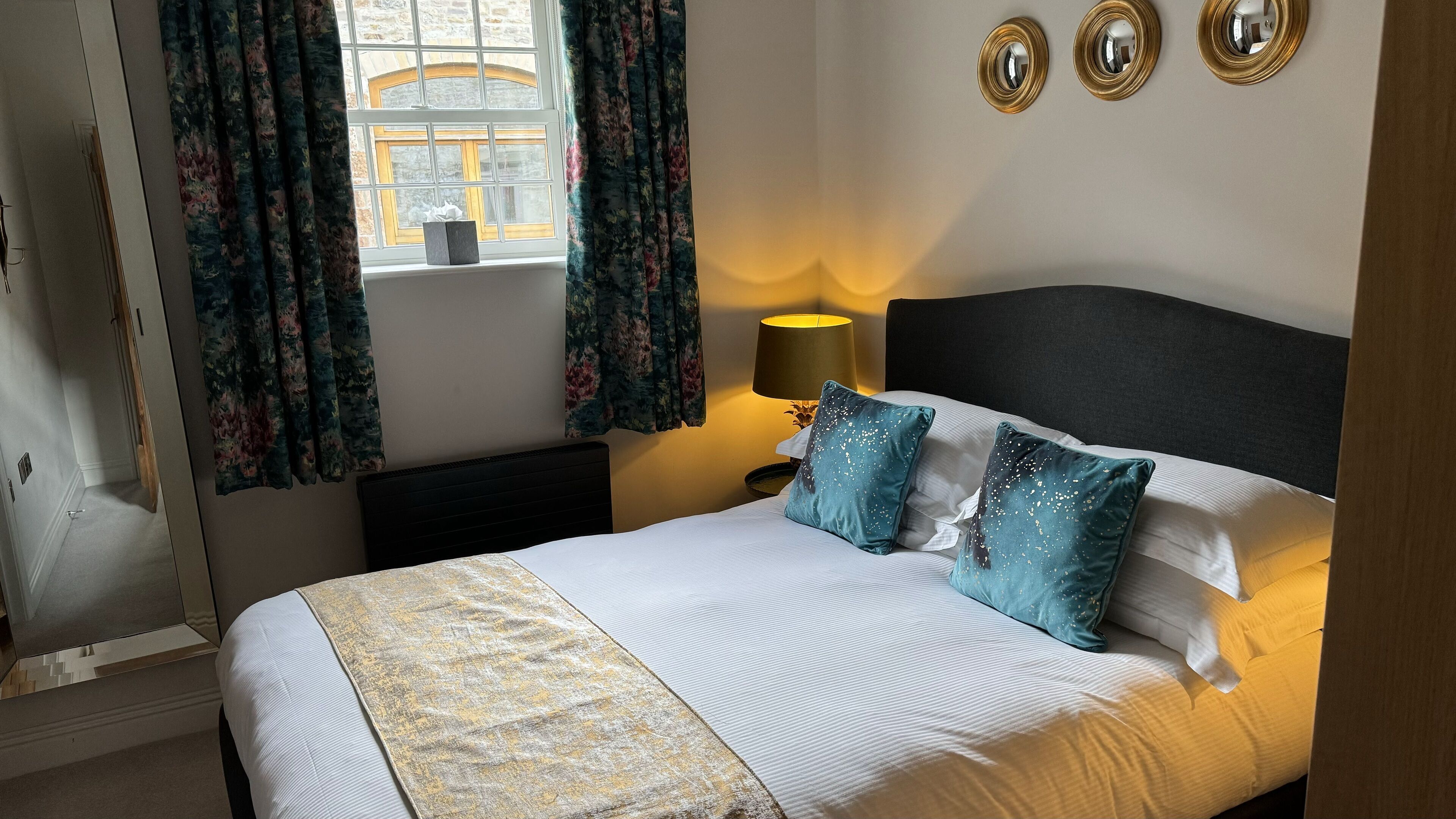 Cama bem arrumada com manta estampada, abajur e dois travesseiros decorativos.
