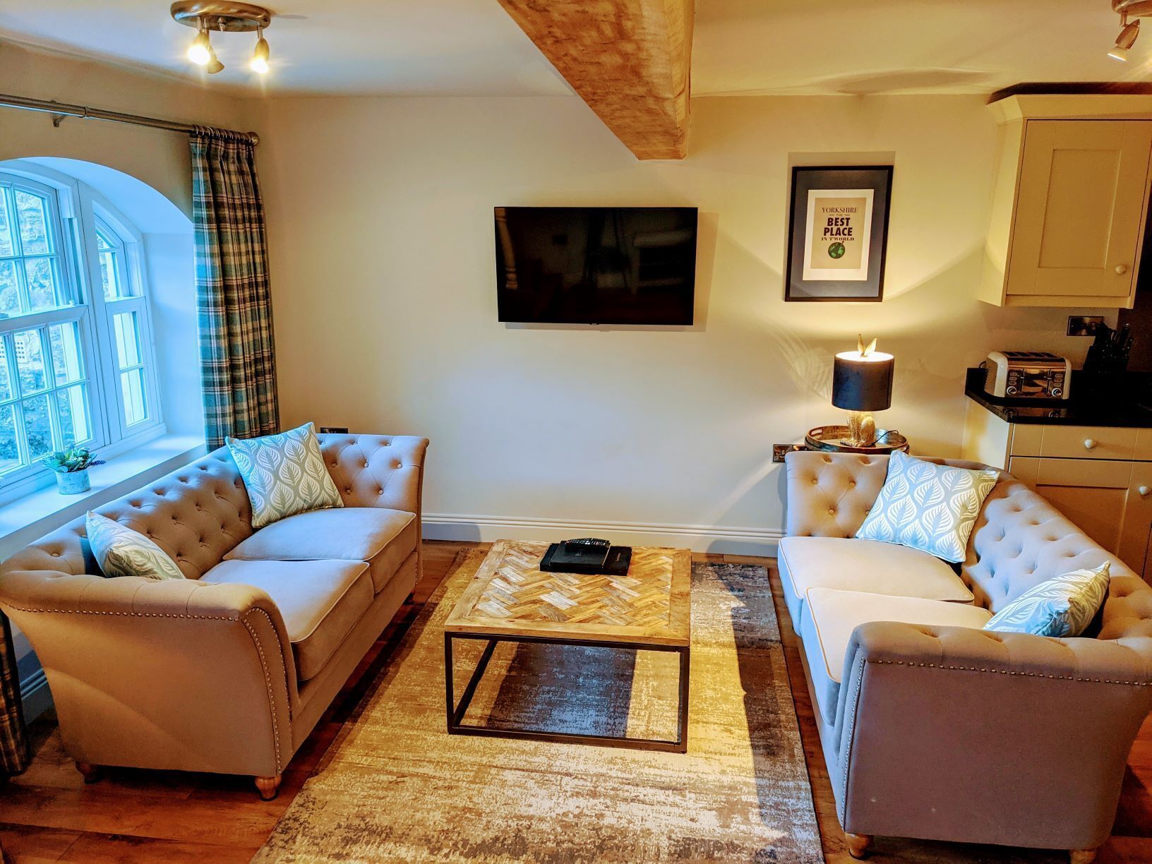 Uma sala de estar acolhedora com dois sofás, uma mesa de centro e uma TV. Há uma janela com cortinas, um quadro emoldurado na parede e uma cozinha ao fundo.