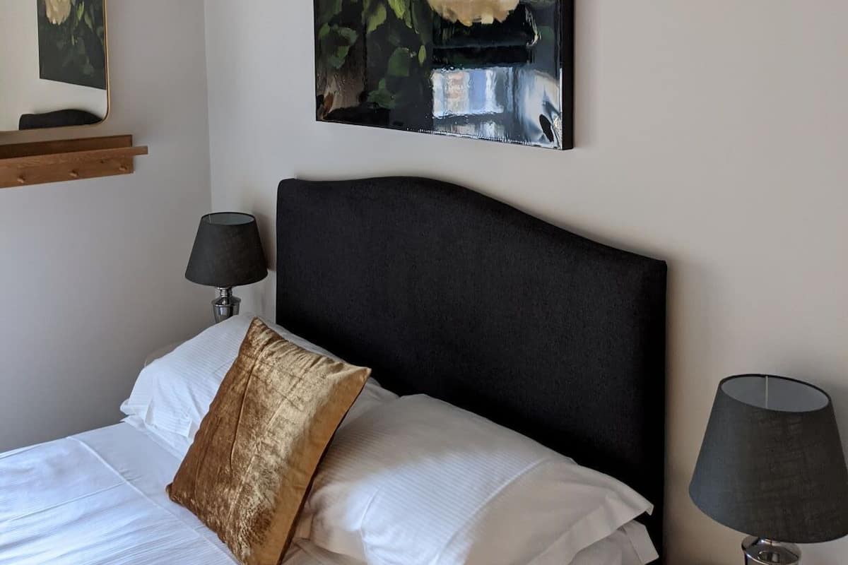 Una cama bien hecha con sábanas blancas y una única almohada decorativa, cabecera negra, mesita de noche con lámpara y un cuadro con flores blancas en la pared.