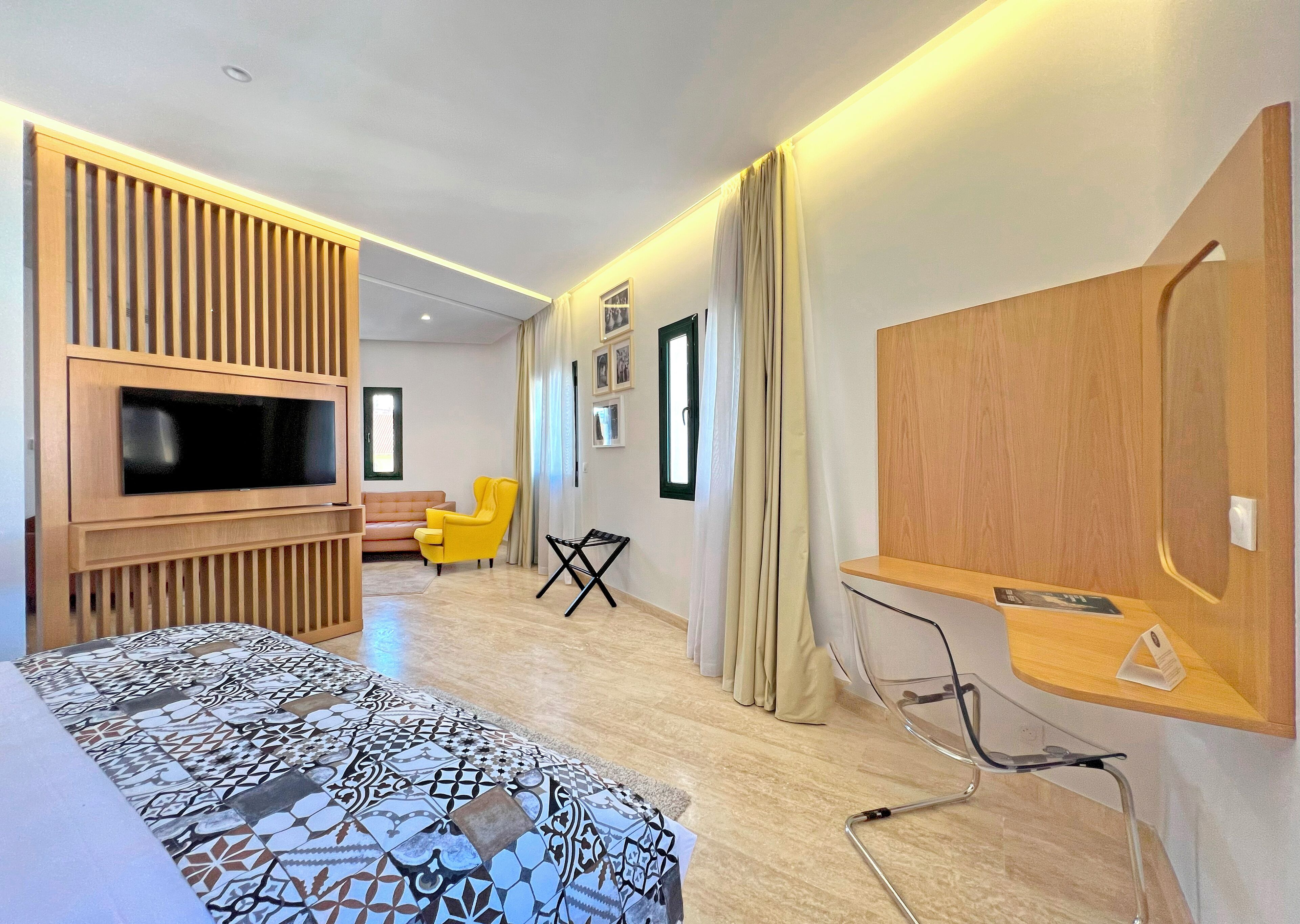 A modern hotel room with a large flat-screen TV, a sofa, a yellow armchair, a desk with a chair, and a patterned rug.