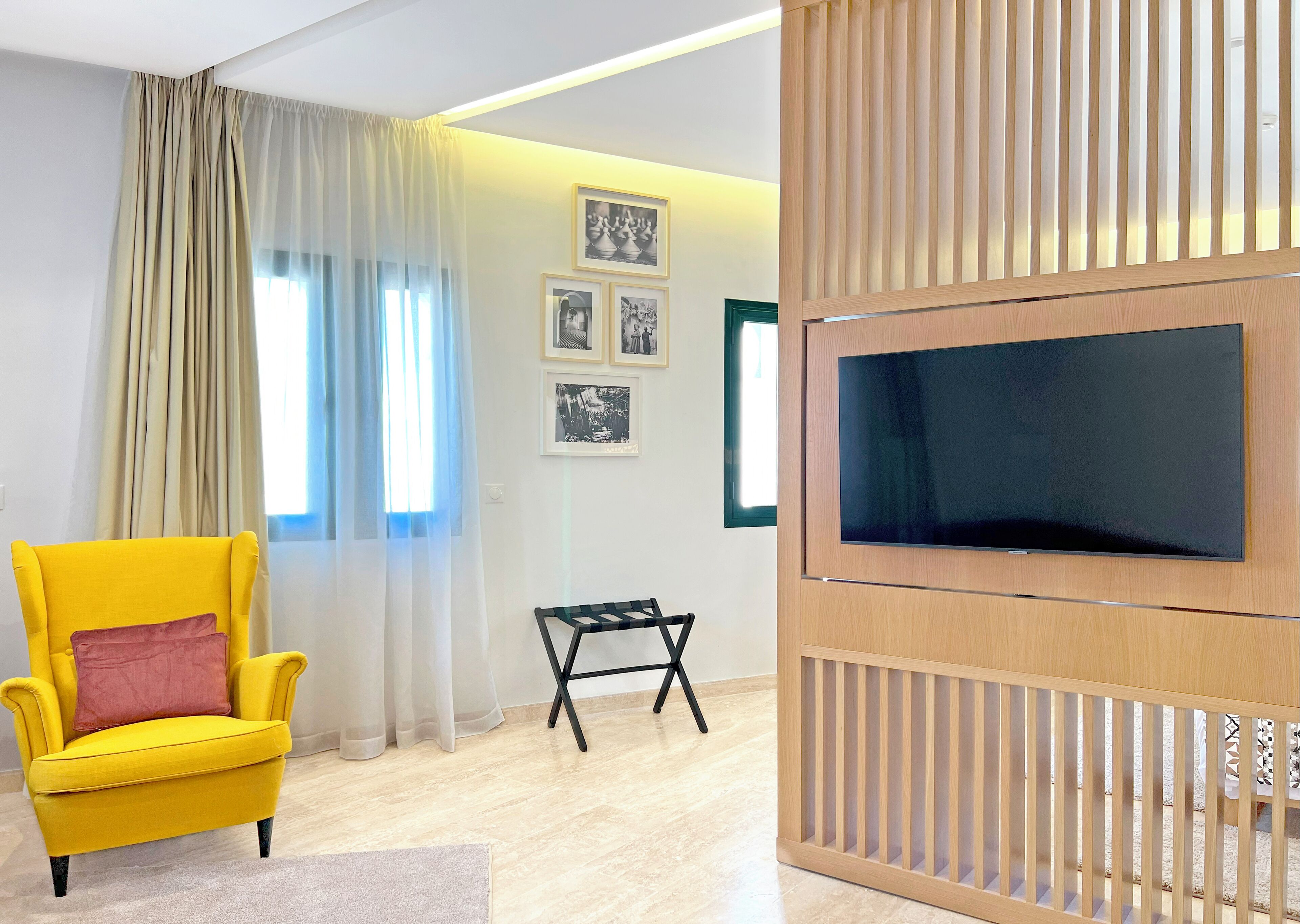 A modern living room with a yellow armchair, a small black table, a TV mounted on a wooden panel, and a window with sheer curtains.