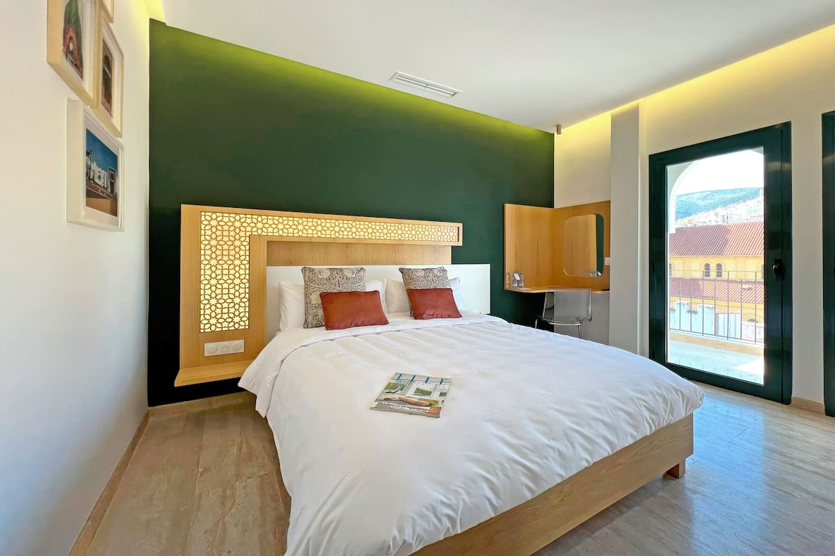 A modern bedroom with a large bed, a wooden headboard, a small desk, and a view of buildings through a glass door.