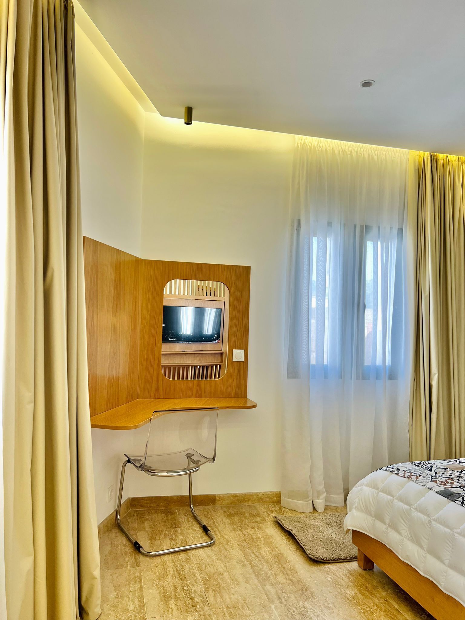 Une chambre d’hôtel avec un meuble en bois pour la télévision, un lit, une chaise et des rideaux en voile.