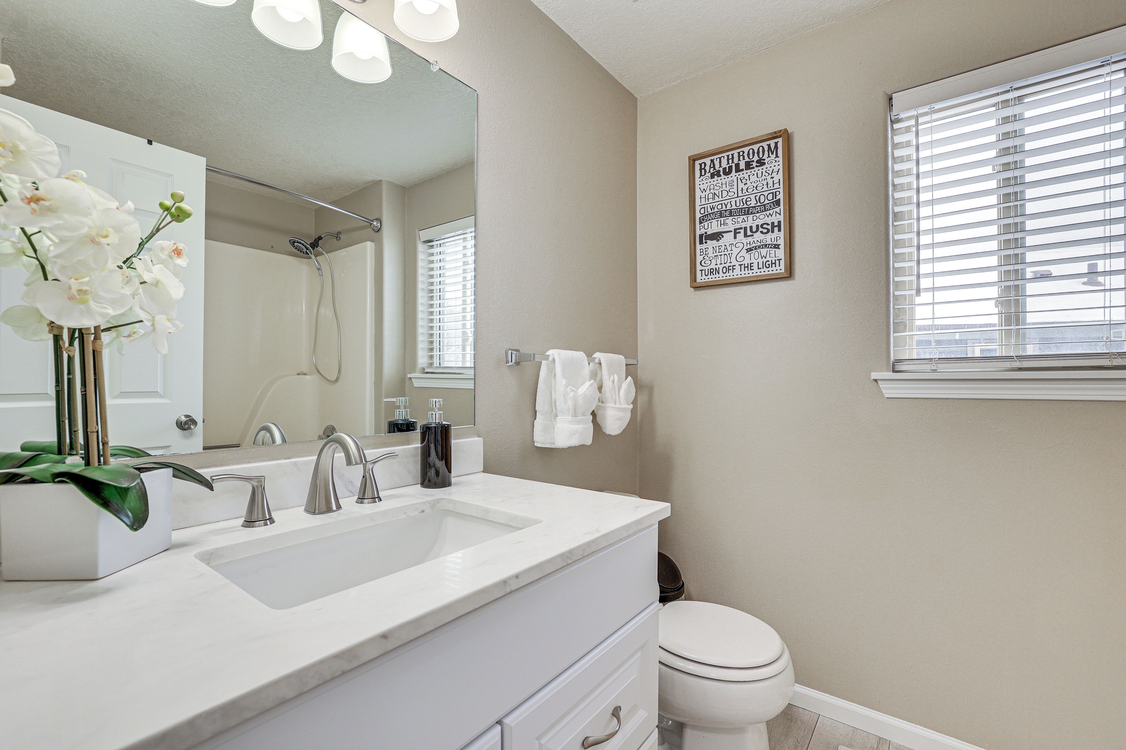 A bathroom with a double sink, a mirror, a shower, and a toilet. There is a sign on the wall and a window with blinds.