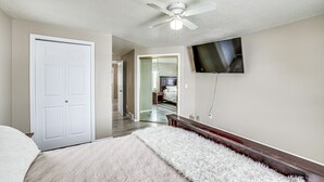 A bedroom with a bed, a ceiling fan, a TV mounted on the wall, and a door.