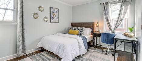 A bedroom with a bed, a desk, a chair, a nightstand, and a window with curtains.
