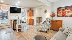 A modern living room with a kitchen area, wooden flooring, a flat-screen TV, and a large floral painting.