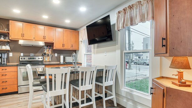 A kitchen with wooden cabinets, a built-in oven, a dining area with white chairs, and a TV mounted above the counter.