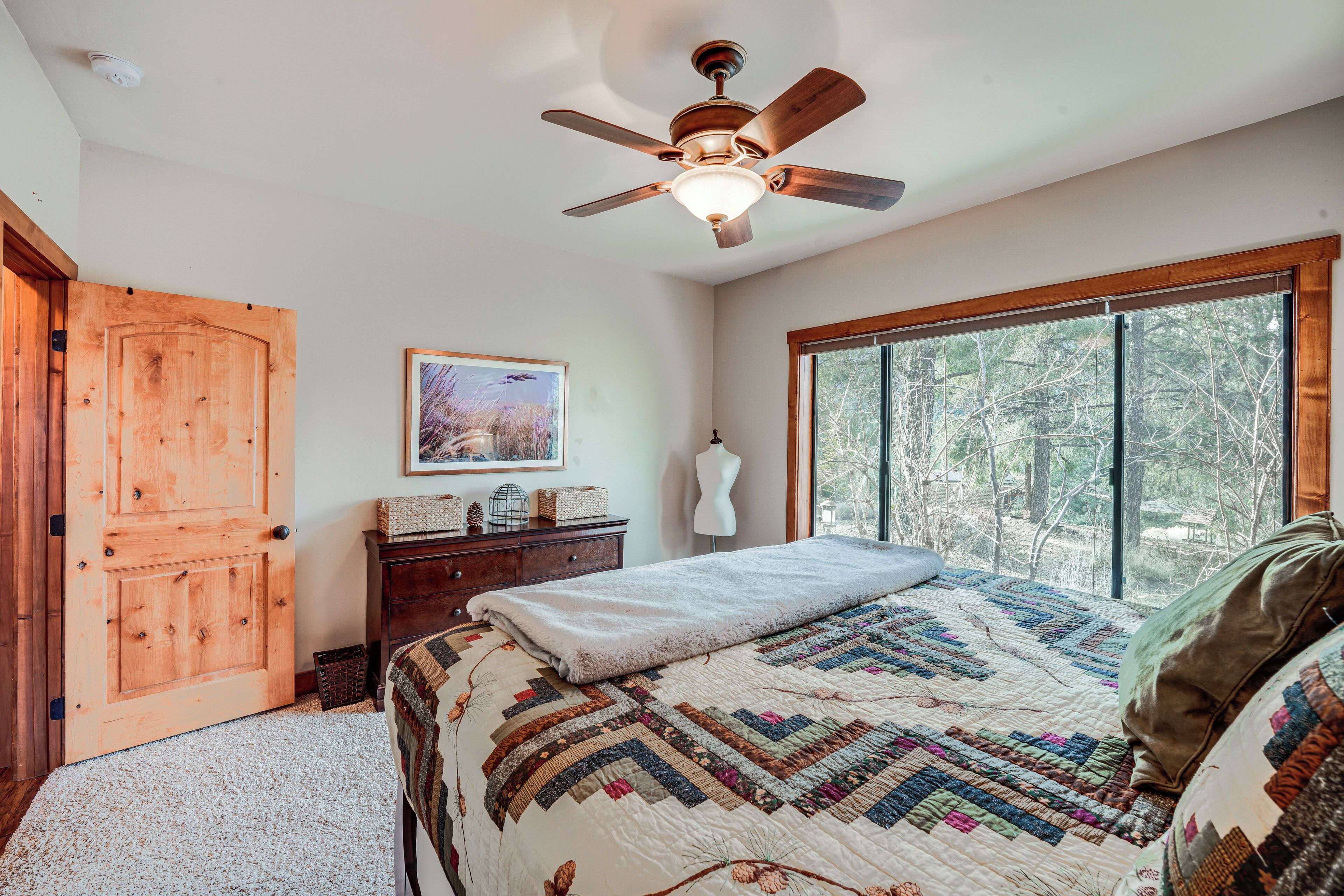 A bedroom with a large bed, a ceiling fan, a window with a forest view, a wooden door, and a dresser with decorative items.