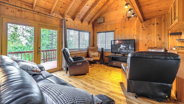 A cozy cabin interior with a wooden ceiling, a large sofa, a recliner, a TV, and a window overlooking greenery.