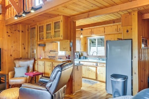 Interior de una cabaña con paredes de madera, una kitchenette, una zona de estar con sofá y sillones, y un cubo de basura.