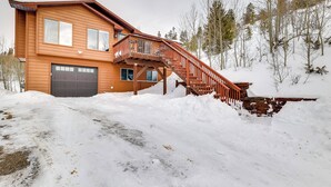 Uma casa de madeira de dois andares com uma escadaria que leva a um deck, cercada por neve e árvores.