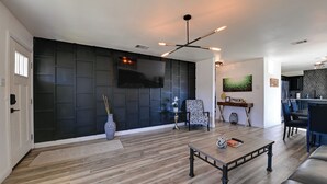 Een moderne woonkamer met een grote flatscreen televisie, donker paneelwerk aan de muur, een eetgedeelte met een bar, en een keuken op de achtergrond.