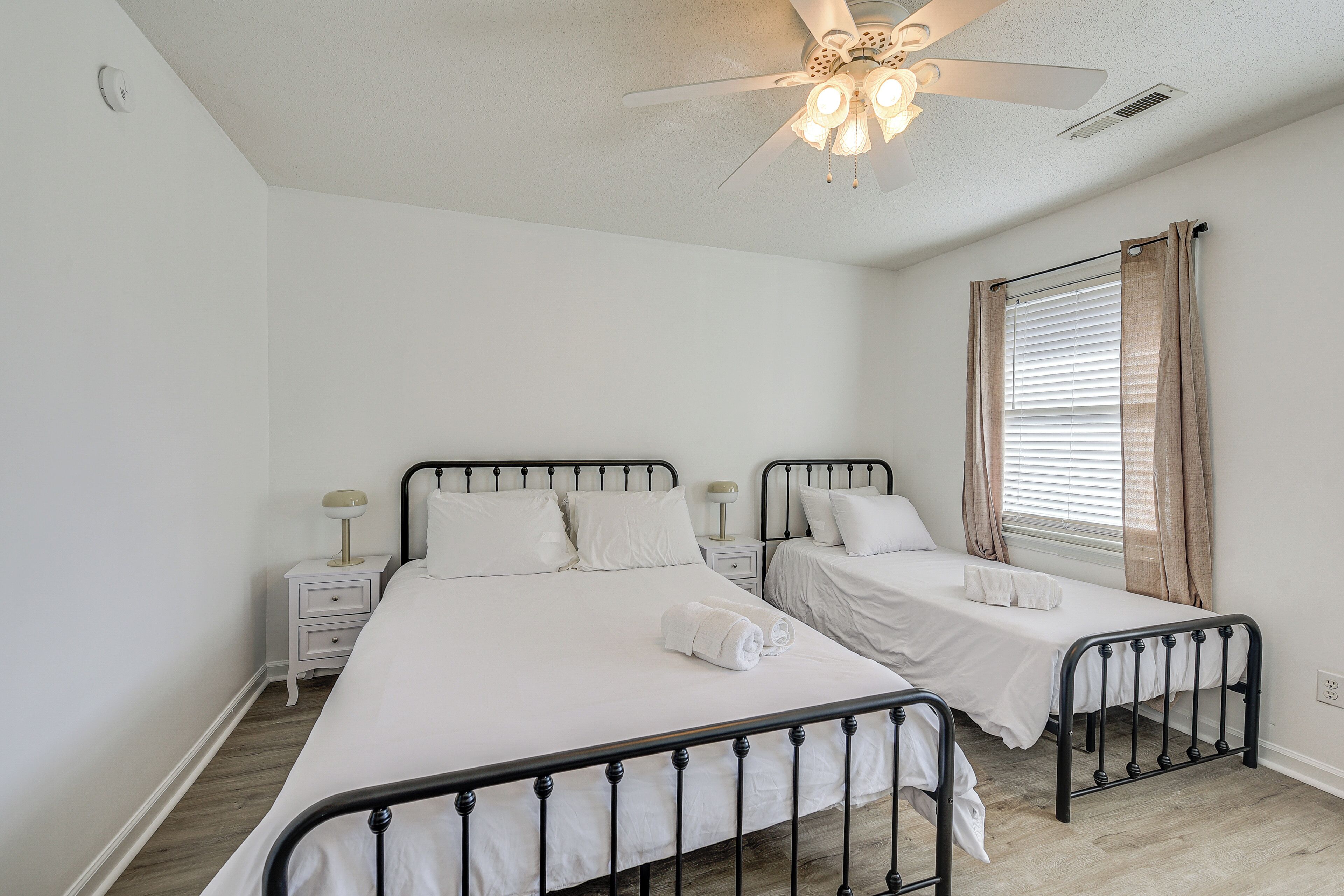 A bedroom with two single beds, a ceiling fan, a window with curtains, and a bedside table with a lamp.
