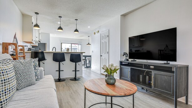 A modern living room with a sofa, a coffee table, and a TV. There is a kitchen area with a large TV and cabinets. - 1 Mi to Beach: Cozy Grand Strand Townhome! (North Myrtle Beach)