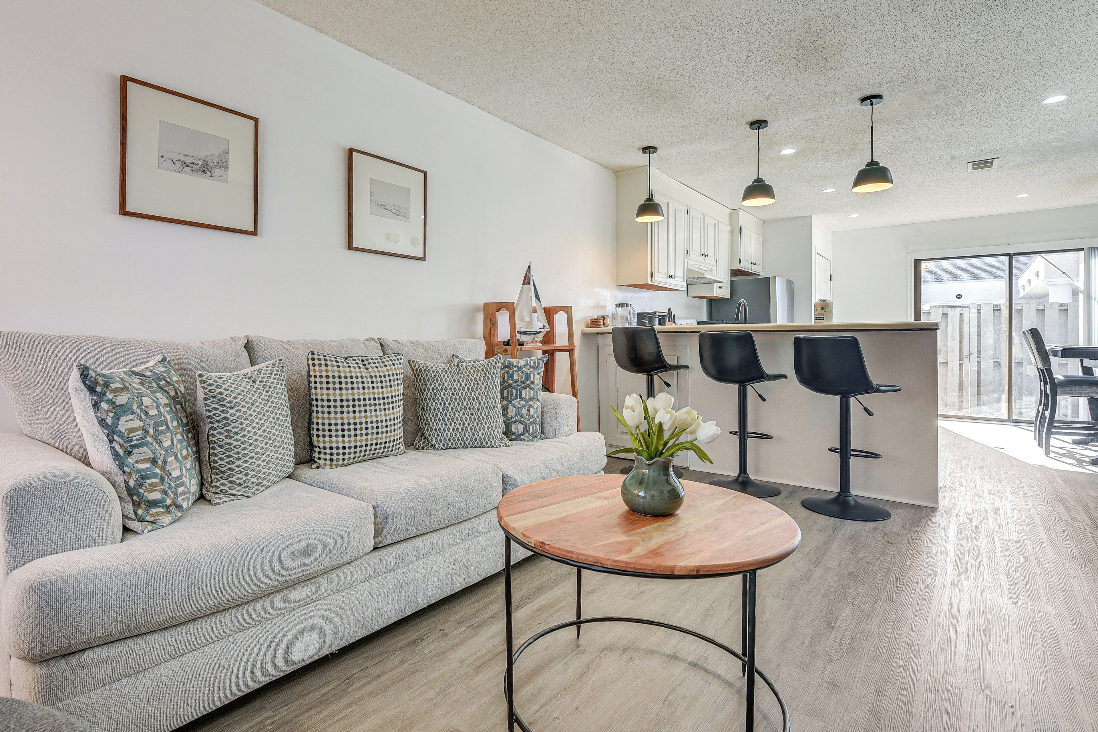 Un salon moderne avec un canapé gris, des coussins à motifs, une table basse en bois et un vase rempli de fleurs blanches. On aperçoit à l’arrière-plan un coin cuisine avec des meubles blancs et des tabourets de bar noirs.