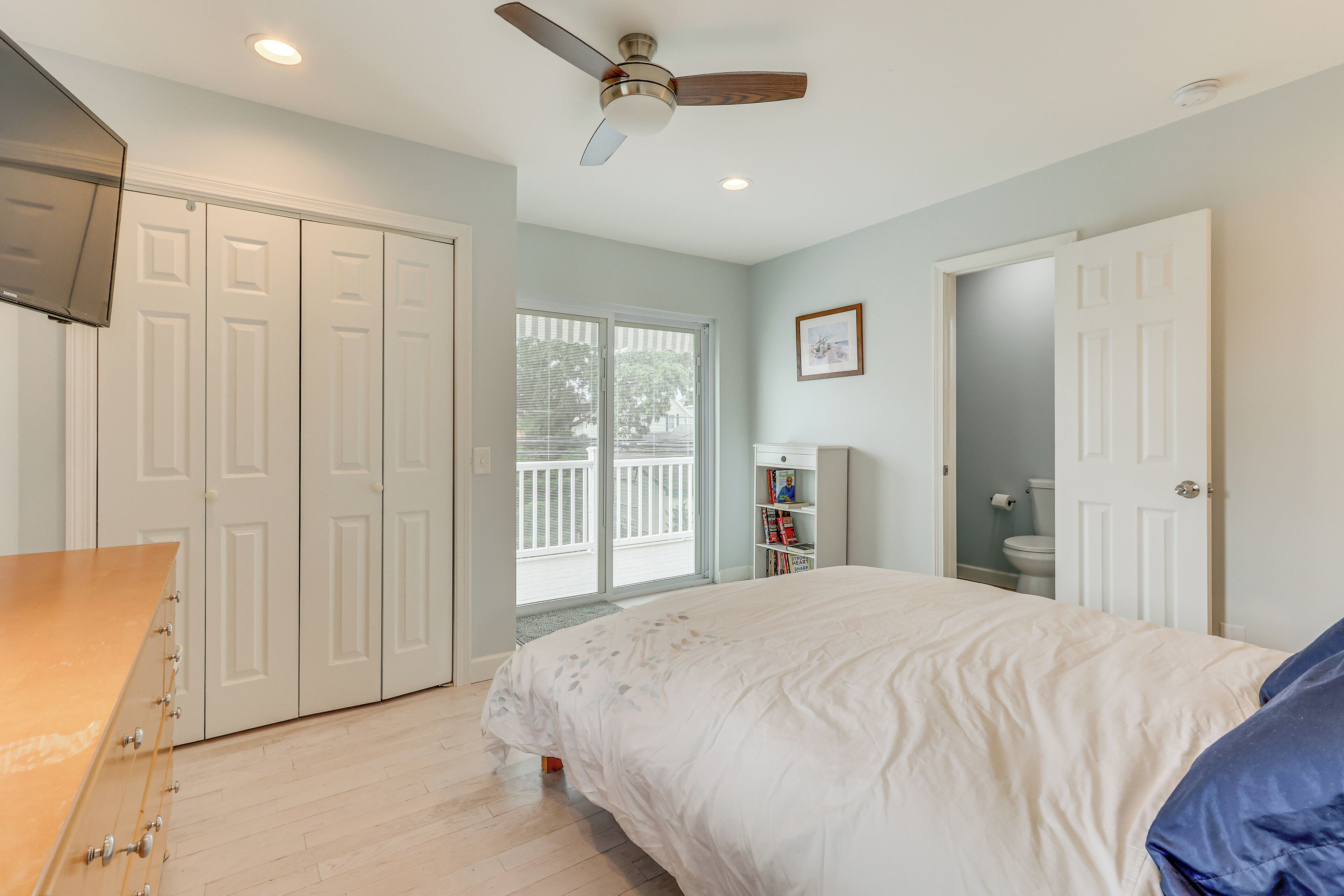 A bedroom with a bed, a ceiling fan, a wardrobe, a window, and a door leading to another room.