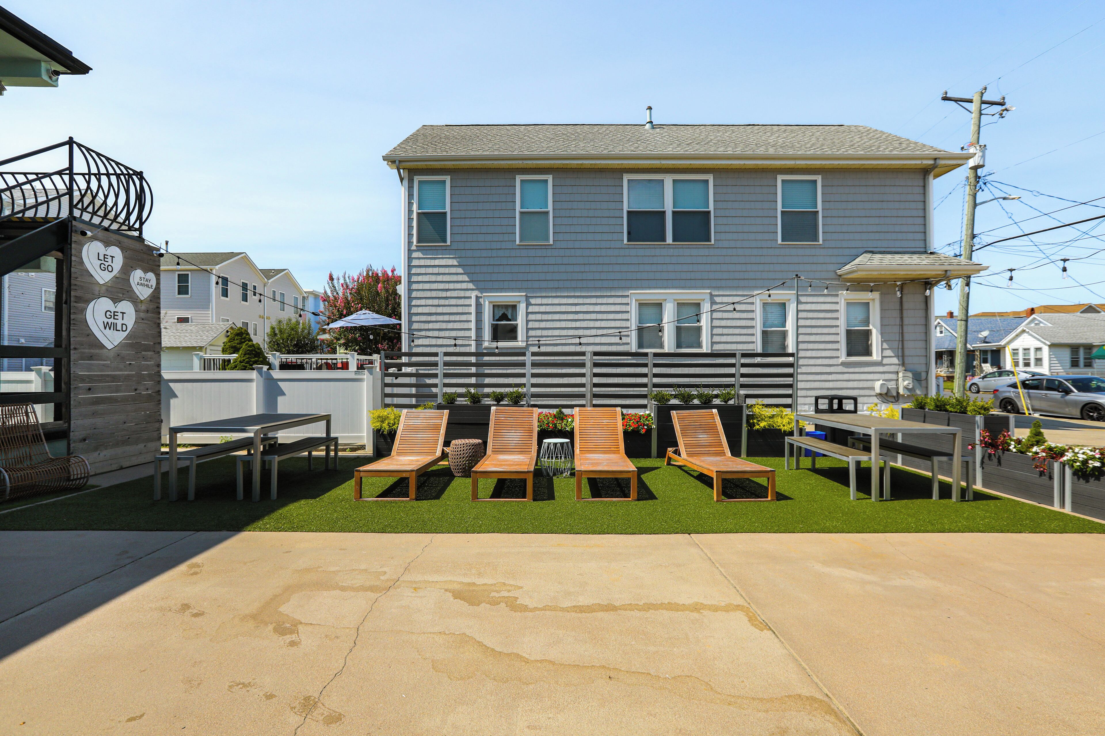A residential building with a front yard featuring four wooden lounge chairs, a concrete patio, and a white fence.