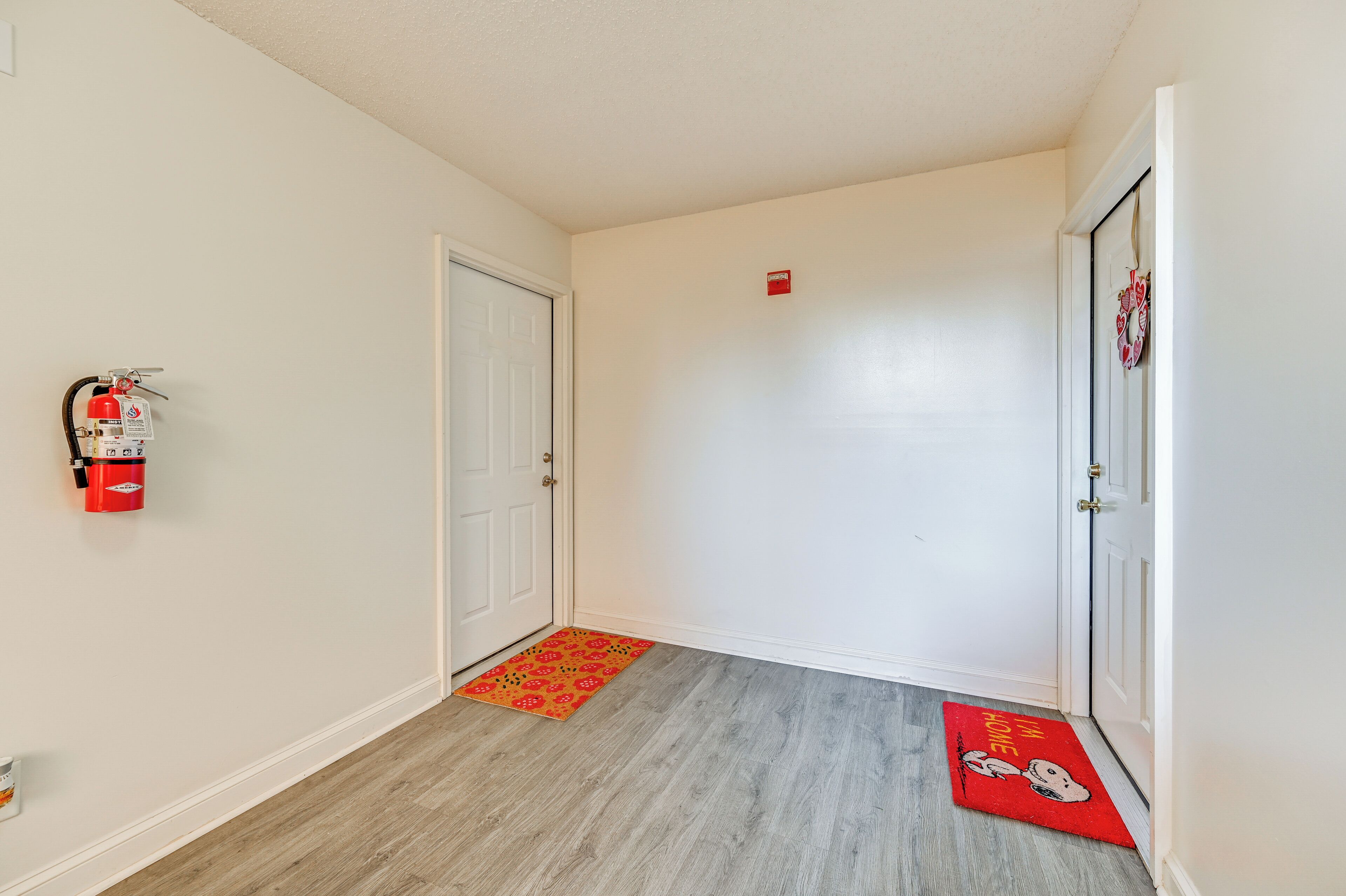 A hallway with a fire extinguisher mounted on the wall, a doormat with a cartoon character, and a door with a small red sign above it.