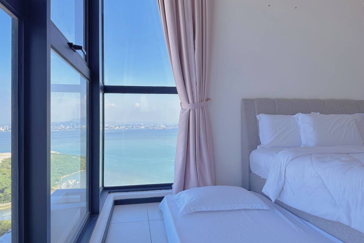 Une chambre avec une grande fenêtre, un lit recouvert de draps blancs, et une vue sur une étendue d’eau et des bâtiments éloignés.