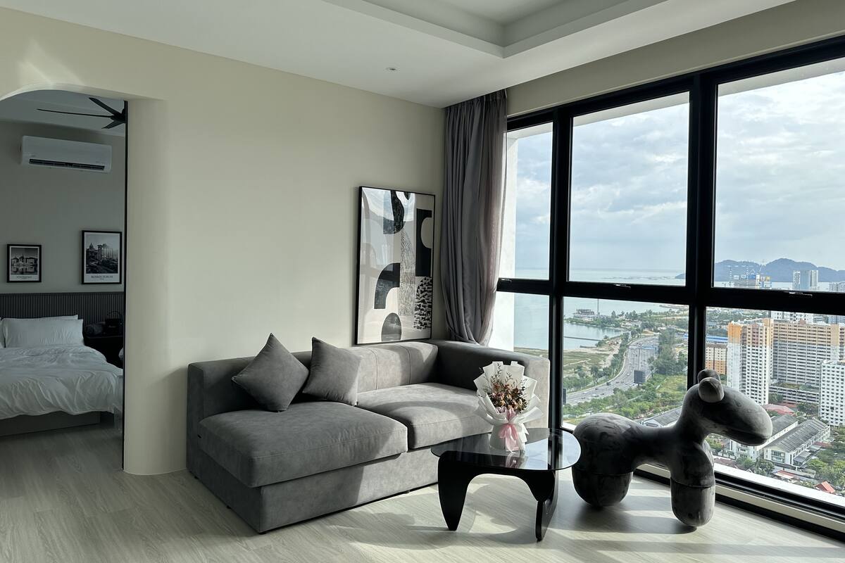 Un salon moderne avec un canapé gris, une table basse en verre et une grande fenêtre donnant sur la ville.