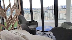 Un salon moderne avec un canapé et deux fauteuils, de grandes fenêtres offrant une vue sur la ville et un panneau décoratif en bois.