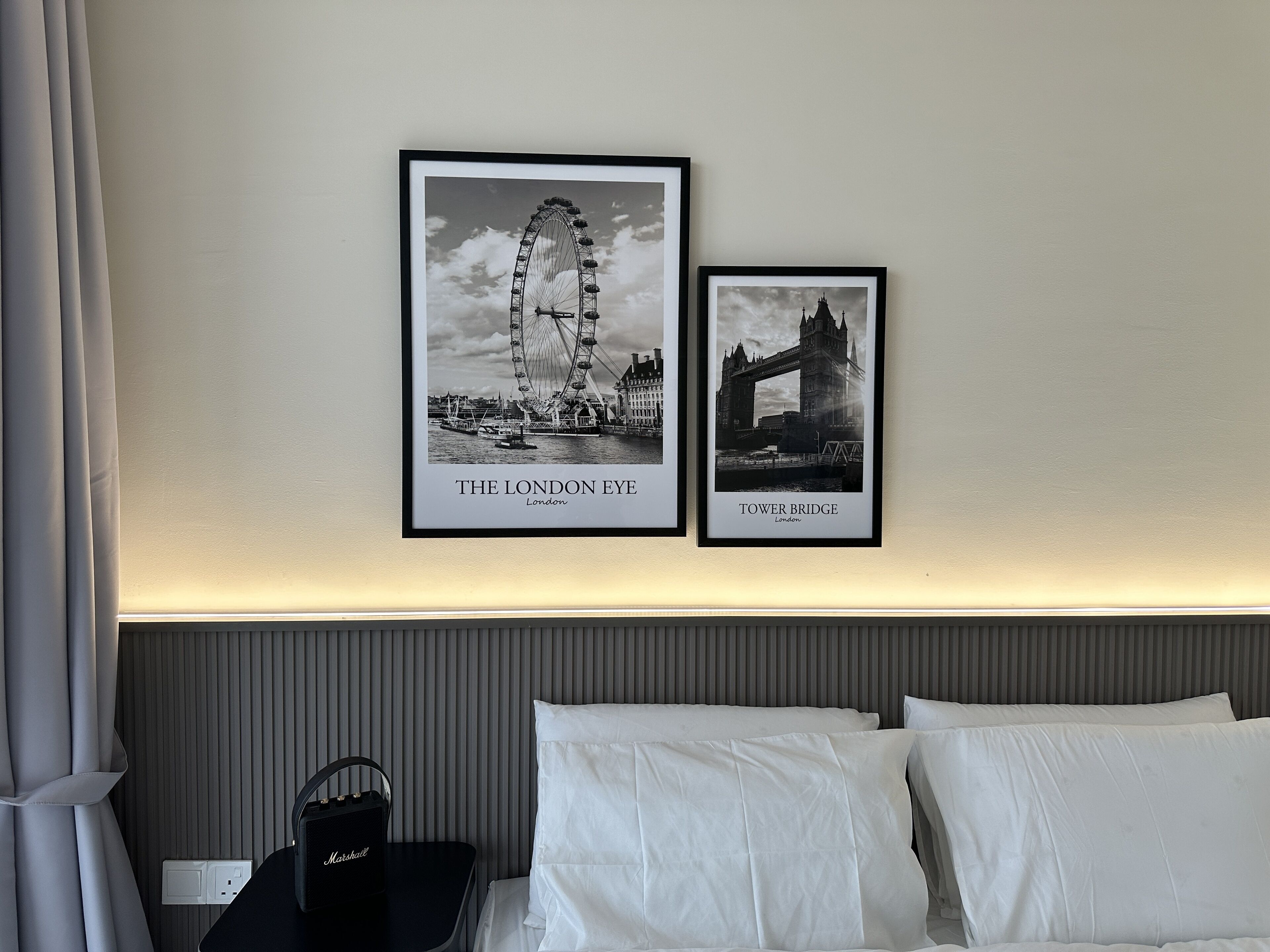 A hotel room with a bed, white pillows, a black nightstand with a lamp, and two framed black and white photographs of London landmarks.