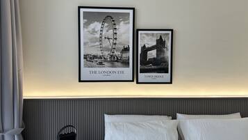 A hotel room with a bed, white pillows, a black nightstand with a lamp, and two framed black and white photographs of London landmarks.