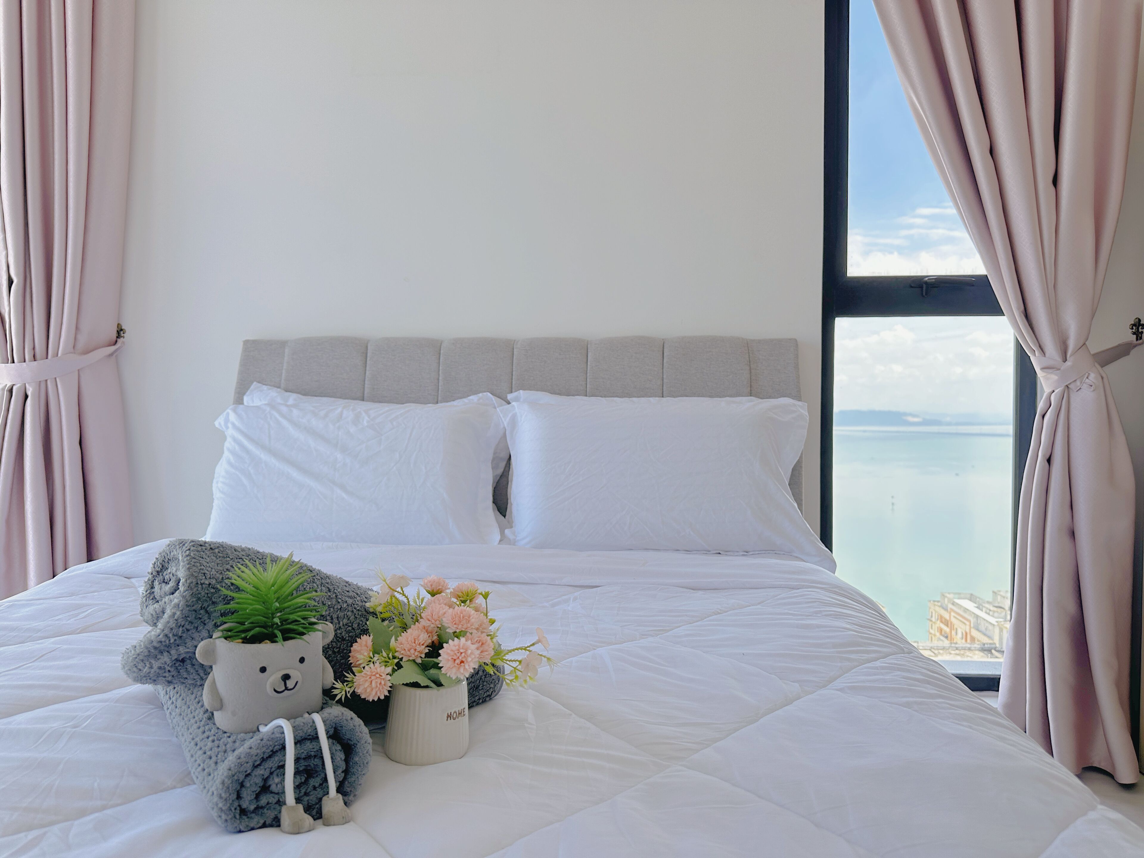 A neatly made bed with white linens, a grey headboard, a plush grey teddy bear, a small potted plant, and pink flowers.