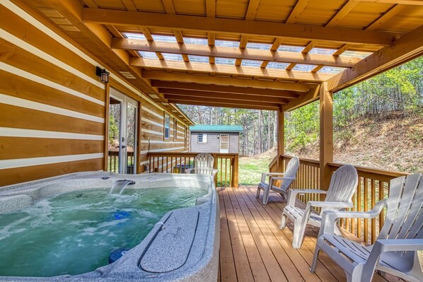 A wooden deck with a hot tub, chairs, and a covered area with a view of the surrounding forest.