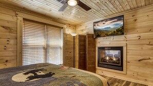 A log cabin bedroom with a fireplace, a large bed, a ceiling fan, and a painting of a landscape. - Hilltop Hideaway 3 Bedroom Cabin by RedAwning (Sevierville)
